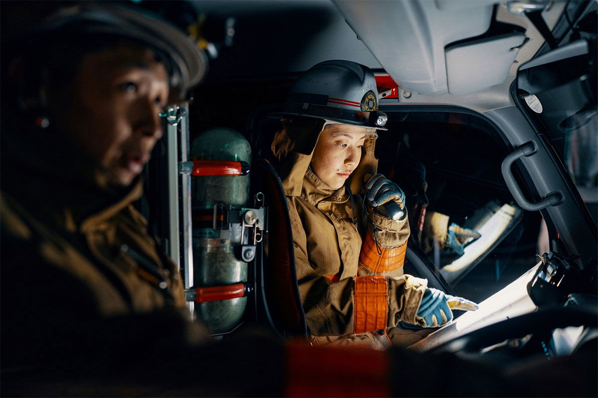 Two firefighters in gear are inside a vehicle. One is speaking into a radio, while the other looks ahead. Emergency equipment surrounds them, and the lighting is dim, suggesting an urgent situation.