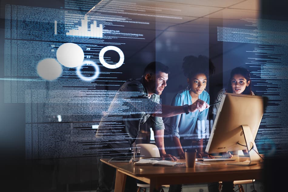 Three people collaborate at a computer desk, with digital data and graphs overlaid, suggesting a tech or programming environment. They seem focused and engaged, discussing or analyzing information on the screen.