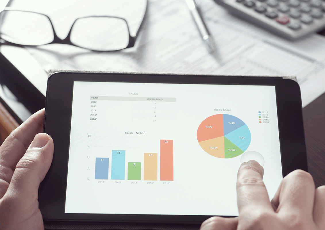 A person uses a tablet displaying financial charts, including a bar graph and a pie chart. The background shows a calculator, glasses, and a document on a desk, suggesting a business or analysis setting.