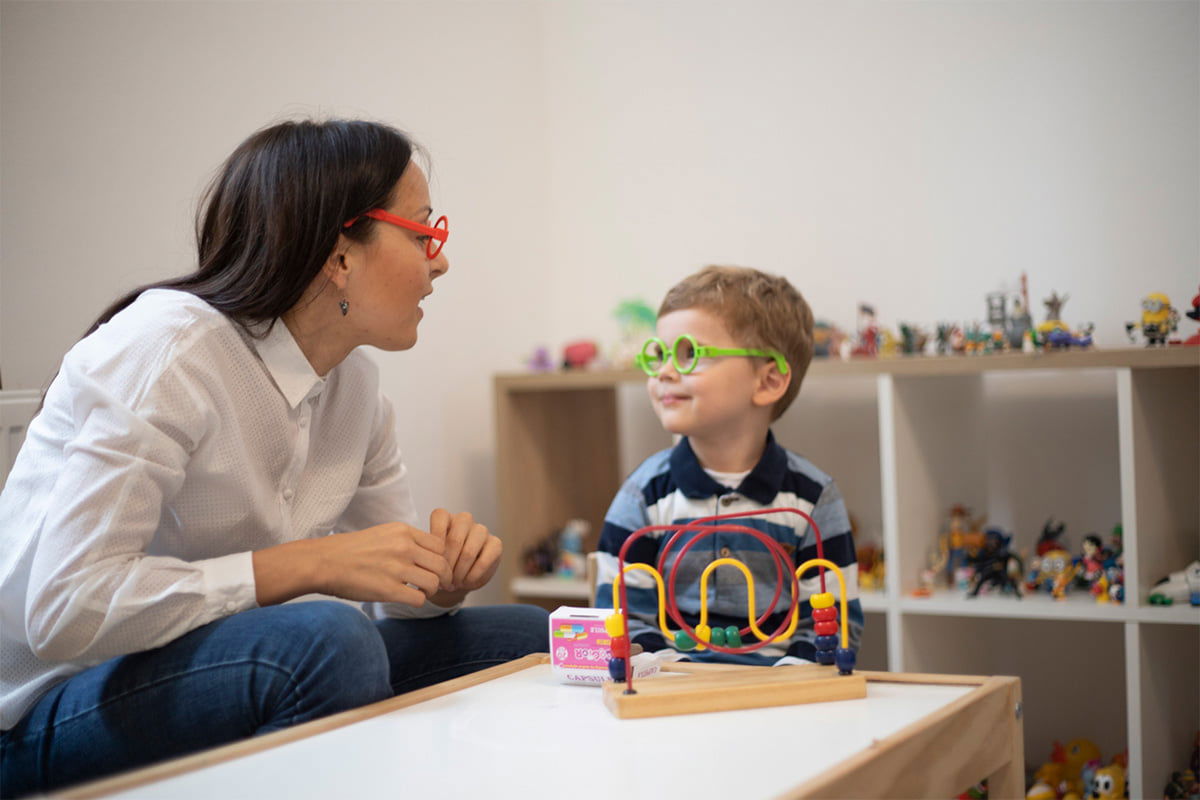 A woman and a child wearing colorful glasses interact happily in a playroom. They are near a toy bead maze and surrounded by various small toys on shelves.