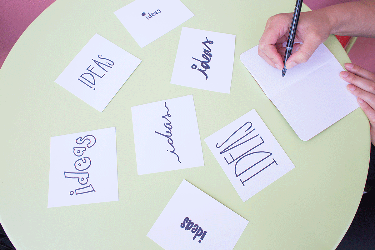 A person writes in a notebook on a round green table. Seven cards with the word "ideas" written in different fonts are scattered on the table.