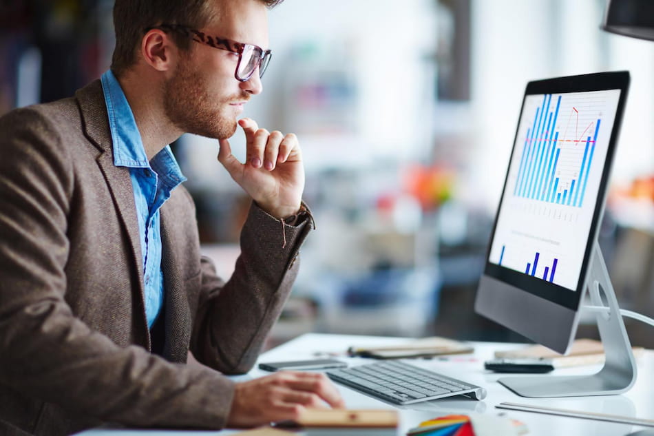 A person in glasses and a brown blazer is seated at a desk, looking intently at a computer screen displaying bar and line graphs. The environment appears to be a modern office setting.
