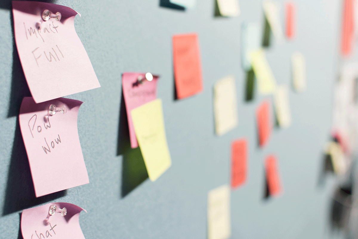 A bulletin board covered in colorful sticky notes with various handwritten words is displayed. The notes are pinned to a blue board, and the focus is on one note that says "Impactful" and "Pow Wow." The background shows more blurred notes.