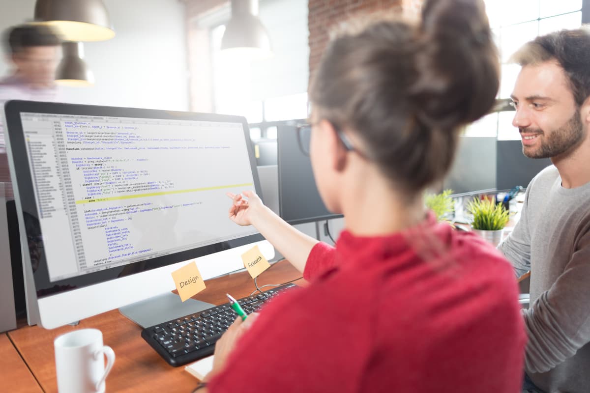 A woman in a red sweater points at a computer screen displaying code, while a man watches attentively. They are in a modern office space with several workstations and plants in the background.