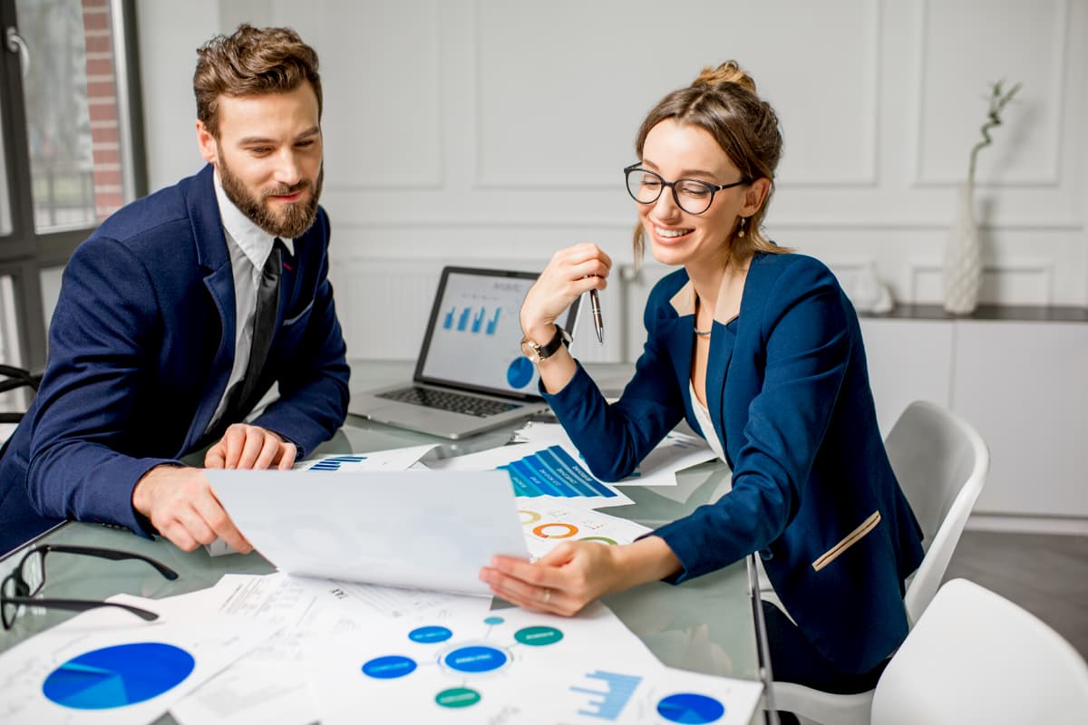A man and woman in business attire are sitting at a table covered with charts and papers, discussing a document. A laptop displaying graphs is open in the background. Both are smiling, suggesting a positive and collaborative meeting.