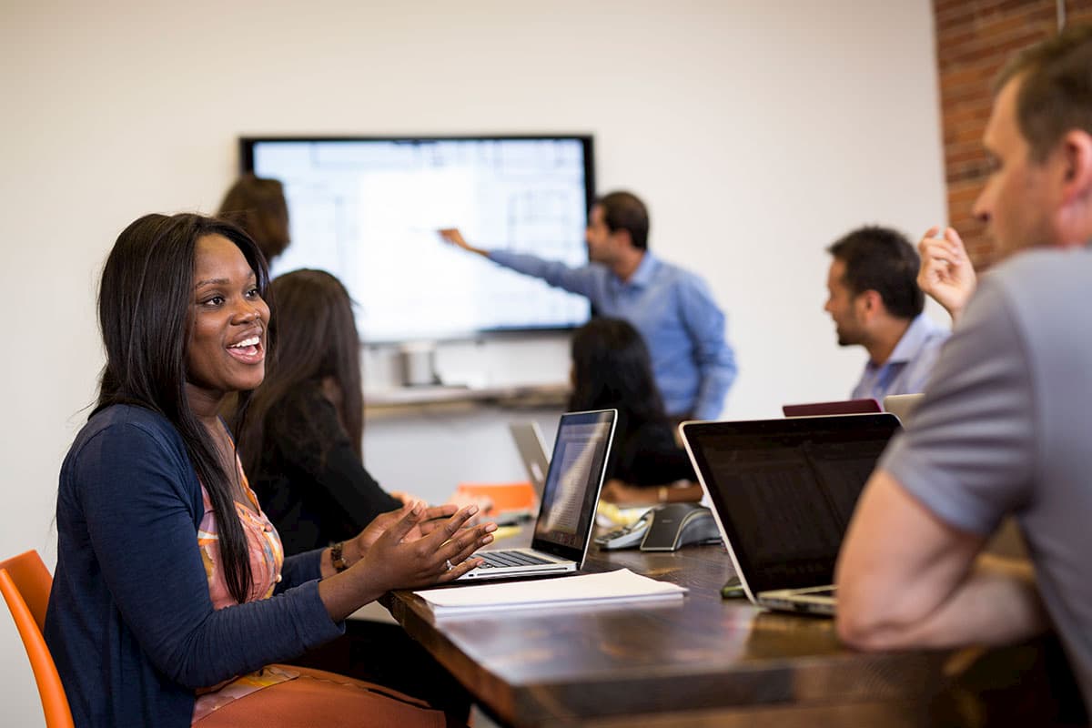 A group of people in a meeting room, seated at a table with laptops. A man stands in the background, pointing at a large screen. A woman in the foreground is smiling, engaged in conversation.