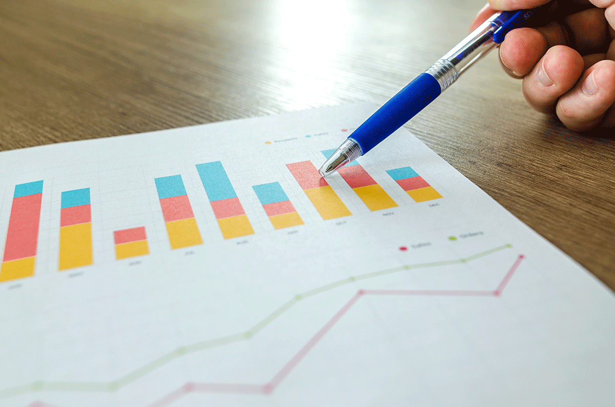 A hand holds a blue pen pointing to a colorful bar chart on a paper. The chart displays various colored bars representing different data categories. Below the bars, a line graph is also visible, all on a wooden table surface.