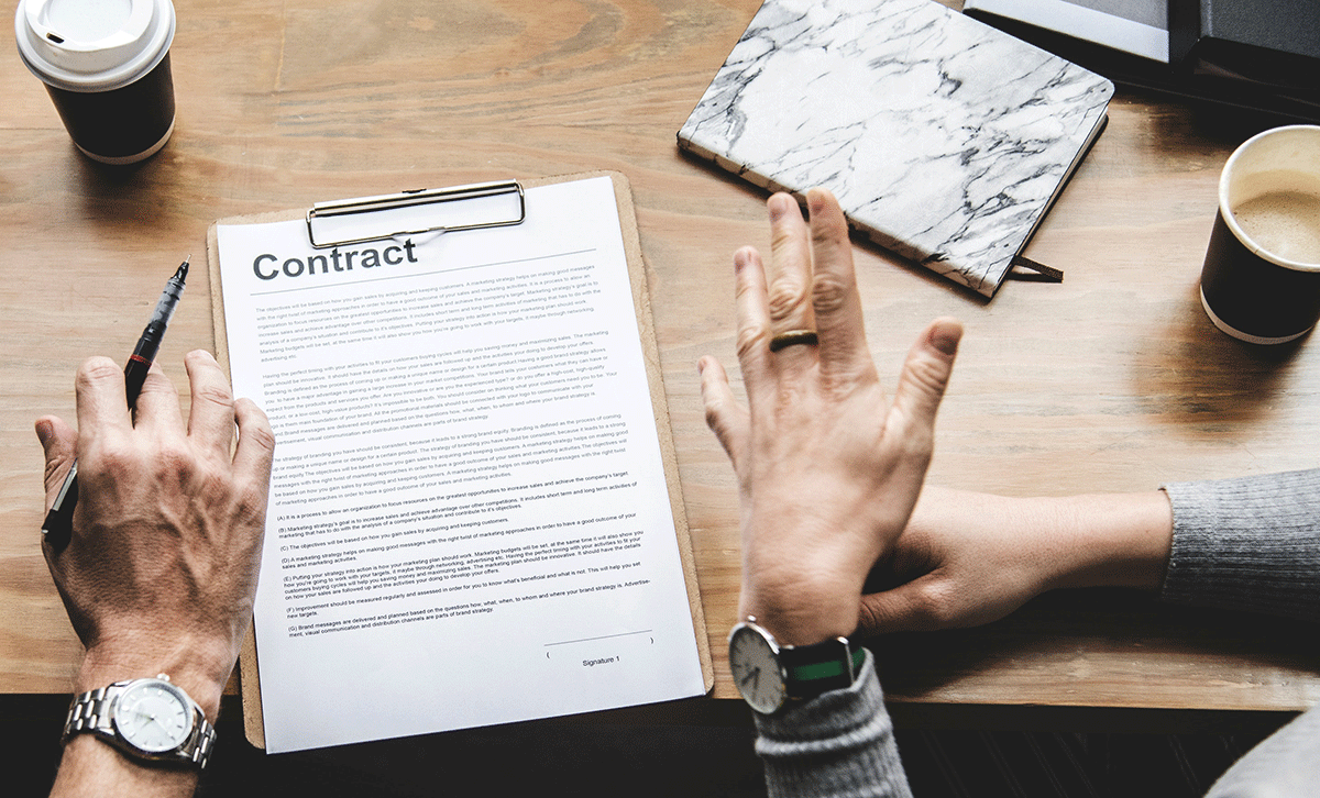 Two people sit at a wooden table with a clipboard holding a contract. One person gestures with their hand, and the other holds a pen. A notebook, laptop, and coffee cups are visible on the table.