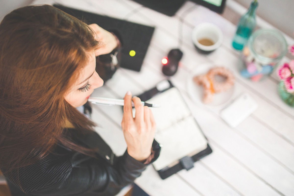 A woman sits at a desk, holding a pen to her lips and looking at an open notebook. Nearby are a smartphone, a cup of coffee, a computer mouse, and a plant. She is wearing a black jacket, and the desk is white.