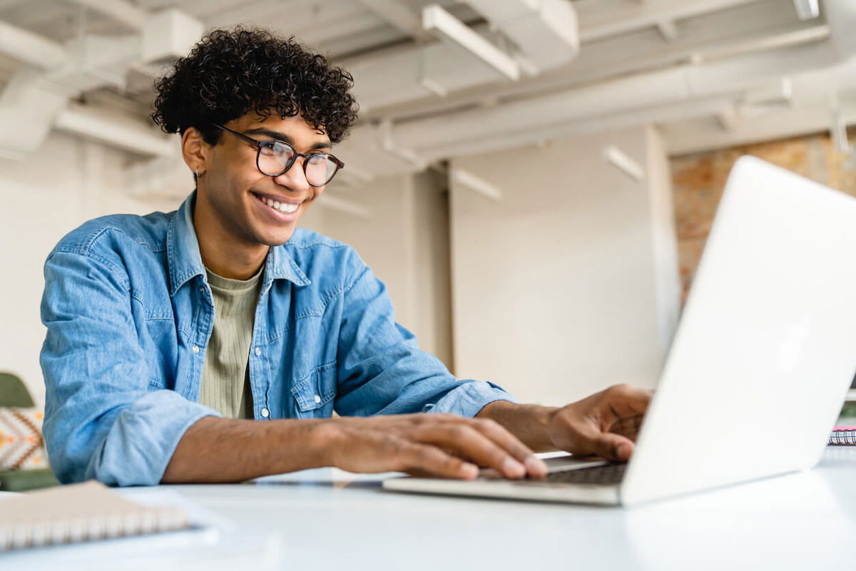 A person with curly hair and glasses is smiling while typing on a laptop. They are sitting at a desk in a bright, open workspace. The individual is wearing a blue shirt over a green top.
