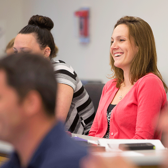 A group of people in a classroom setting, with a woman in a pink cardigan smiling and looking engaged. Others are seated nearby, focusing on what's happening in the room.