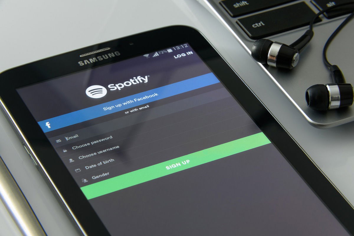A smartphone displays the Spotify sign-up screen with options to sign up via Facebook or email. The phone is placed on a desk next to a laptop and a pair of earbuds.