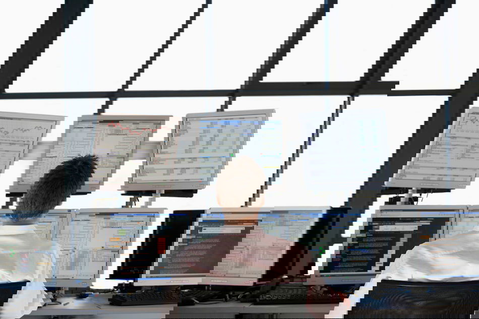 A person is sitting in front of multiple computer monitors displaying financial data and graphs. The room has large windows, allowing natural light to fill the space. The individual appears to be analyzing the information on the screens.
