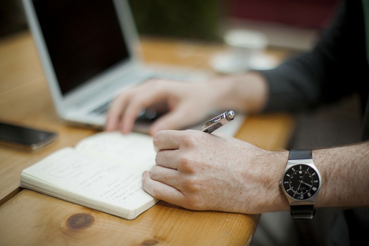 A person is sitting at a wooden table, writing in a notebook with a pen. A laptop is open nearby, along with a smartphone and a coffee cup, creating a workspace environment. The person is wearing a watch on their left wrist.