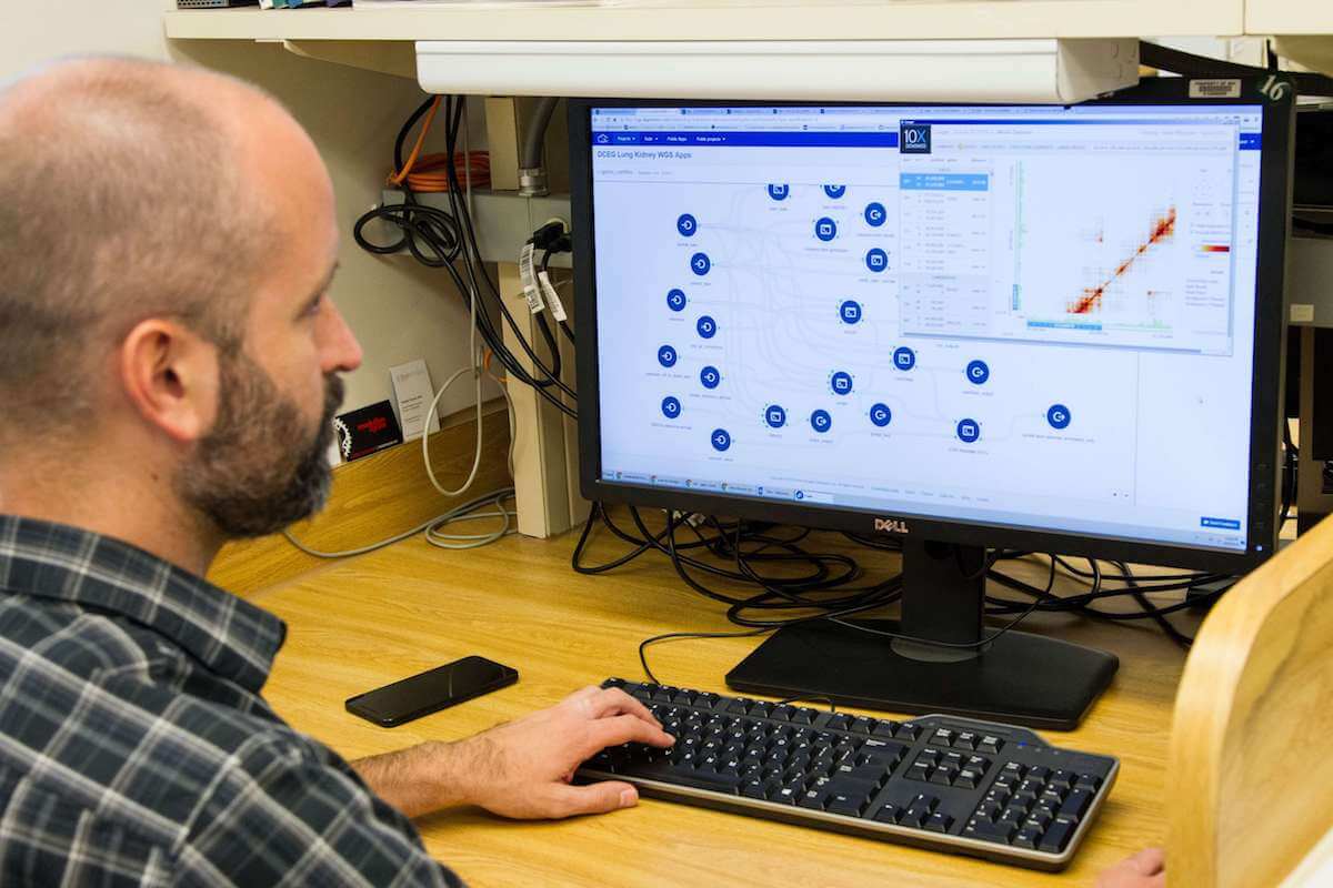 A person sits at a desk looking at a computer screen displaying a network diagram. Various nodes and connections are visible on the monitor. A phone and a piece of paper are on the wooden desk.