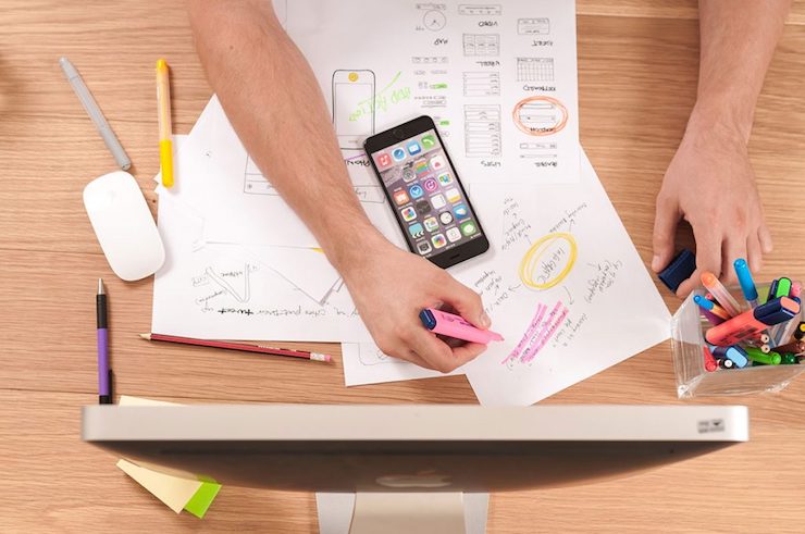 Person working at a desk with scattered papers, a smartphone, and a desktop computer. They're holding a pink highlighter, marking text. A container of colorful markers is nearby.
