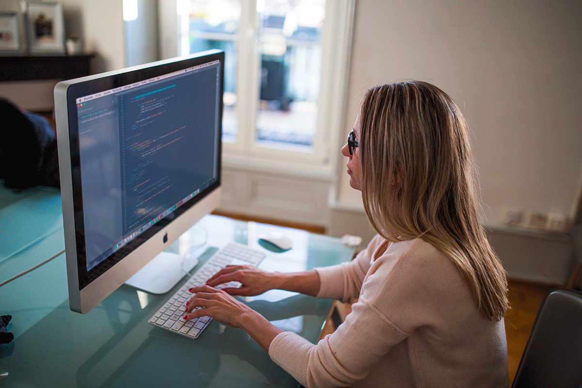A woman with long blond hair, wearing glasses, is working on a computer. She is typing with both hands on a keyboard in front of a large monitor displaying code. She is seated at a desk near a window with sunlight streaming in.