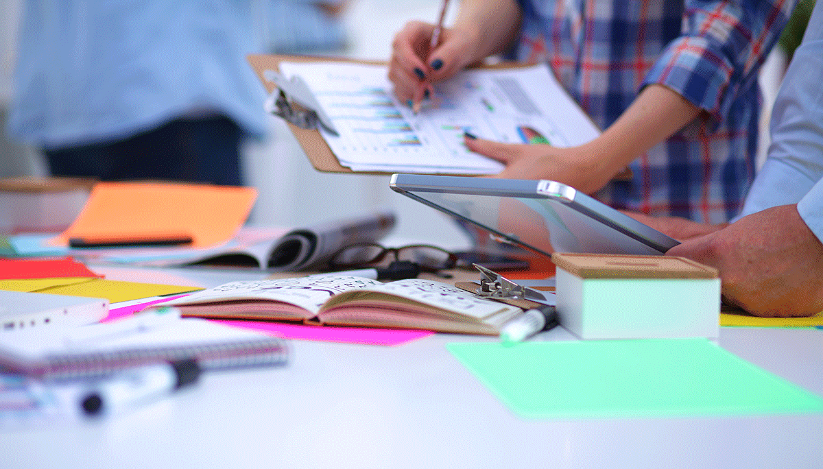 A group of people working at a desk, using tablets and reviewing charts on a clipboard. The desk is cluttered with colorful sticky notes, notebooks, and folders, creating a busy and collaborative office atmosphere.