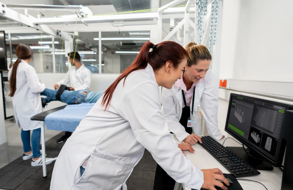 Two medical professionals in lab coats work at a computer analyzing data, while two other people in lab coats attend to a person lying on a table in the background. The room appears to be a medical laboratory or clinic.