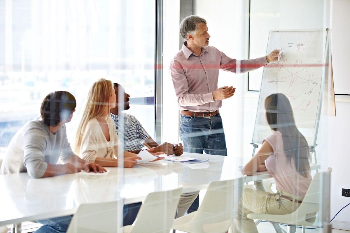 A man presents at a whiteboard in a modern conference room, while four colleagues sit at a table and listen attentively. The scene is bright, with large windows in the background.