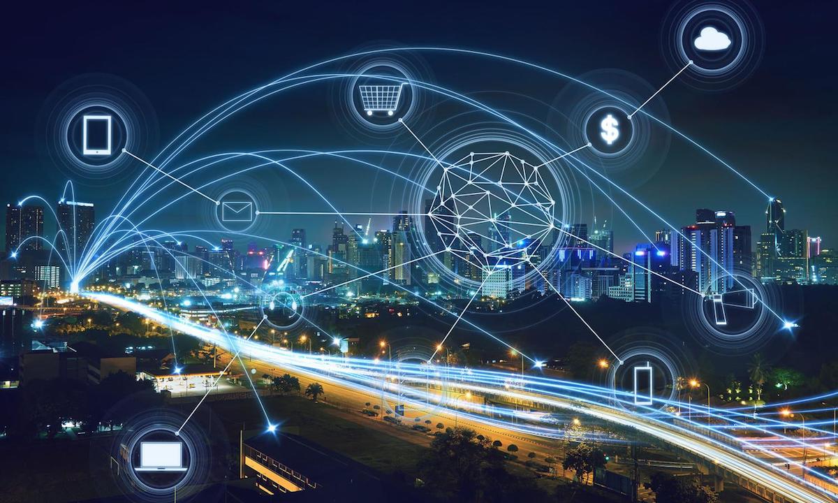 A cityscape at night with glowing lines and connected icons representing a digital network. The icons include a shopping cart, cloud, dollar sign, and smartphone, symbolizing technology and connectivity in an urban environment.