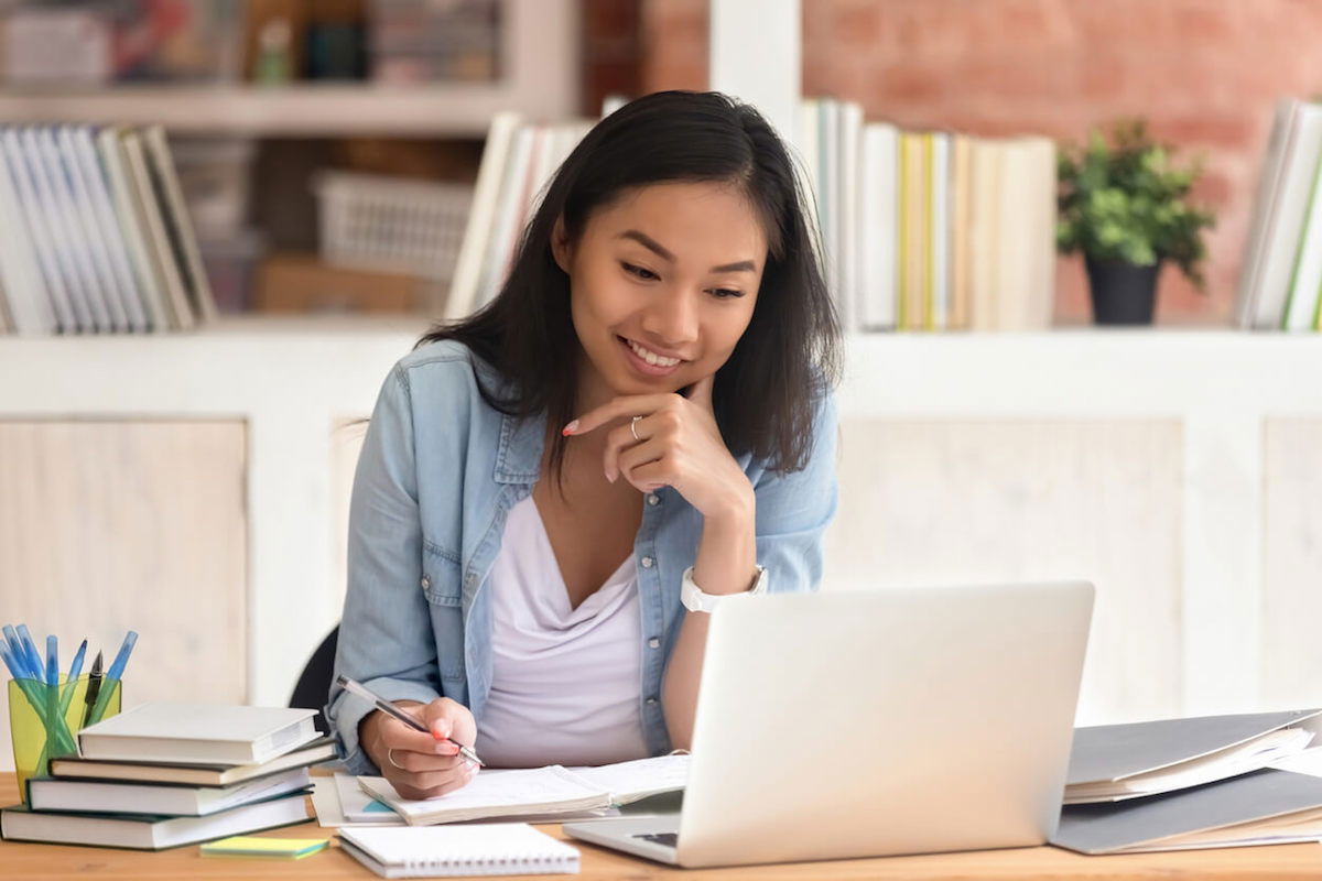 A woman sits at a desk with an open laptop, smiling thoughtfully. She holds a pen and is surrounded by books and notebooks. A shelf with books and a potted plant is in the background.