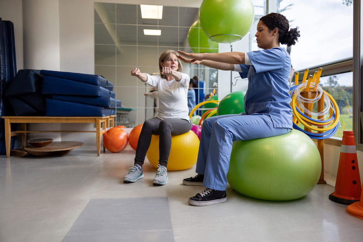 A woman in casual attire sits on an exercise ball, guided by a healthcare professional in scrubs, in a therapy room with bright exercise balls and equipment. They perform arm stretches, focusing on rehabilitation or fitness.