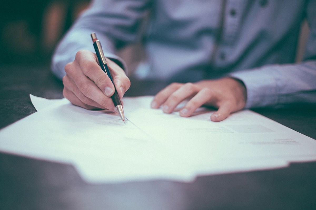 A person in a blue shirt is holding a pen and writing on several sheets of paper on a desk. The background is blurred, emphasizing the hand and pen.