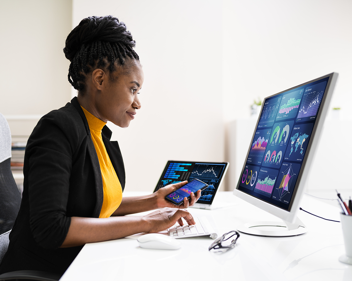 A person in a yellow top and black blazer examines data on a large computer screen displaying graphs and charts. They hold a phone in one hand and use a keyboard with the other. A laptop with similar data is open nearby on a white desk.