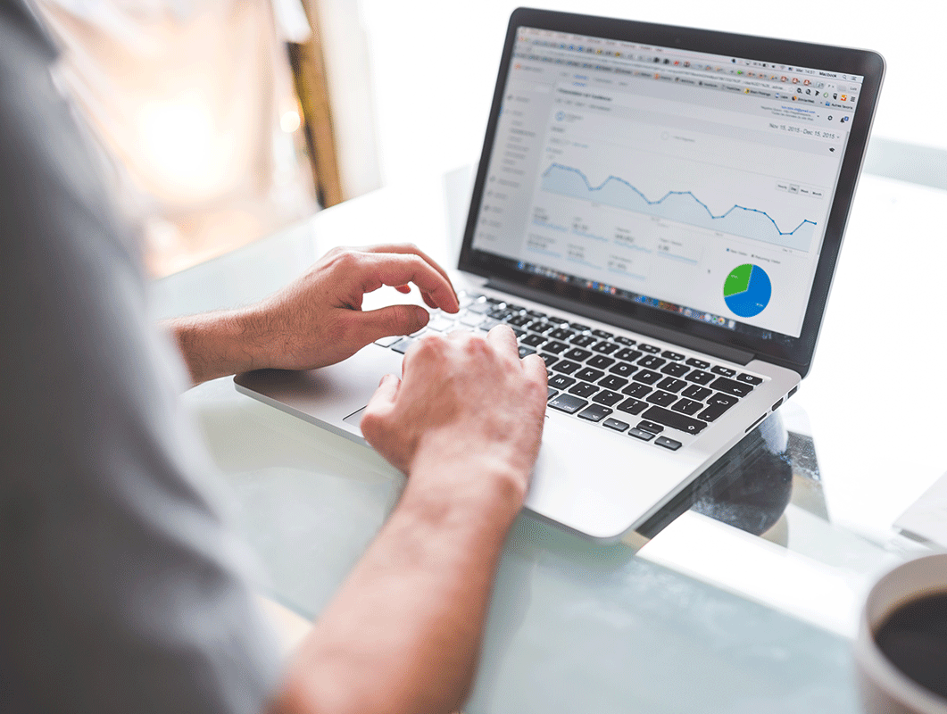 A person types on a laptop displaying analytics, including line graphs and a pie chart, indicating data trends. The laptop is on a glass table with a coffee mug nearby, suggesting a work setting.