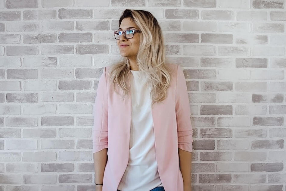A person with long hair, wearing glasses and a pink blazer over a white top, stands against a white brick wall, looking to the side.