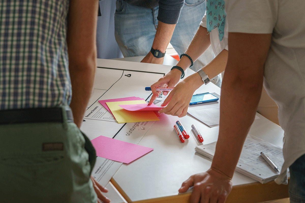 A group of people collaborates at a table with notebooks, colorful sticky notes, pens, and a smartphone. One person is pointing at the notes, while others are leaning in, suggesting a brainstorming session or team discussion.