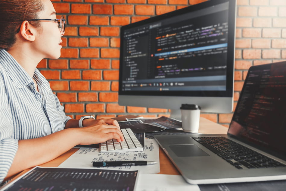 A person wearing glasses is focused on computer screens displaying code. They are sitting at a desk with a keyboard, coffee cup, and papers. The background features a brick wall, creating a tech-oriented workspace atmosphere.