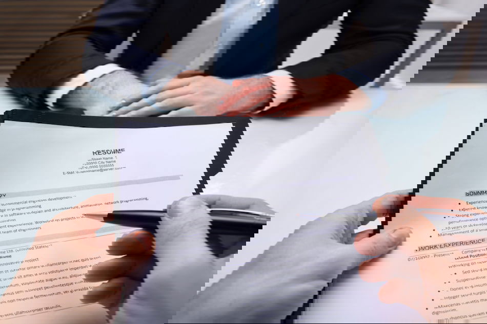 A person across a table is holding a pen and reviewing a resume. The document highlights work experience and skills. Another person, wearing a suit, sits opposite with hands folded, suggesting a job interview setting.