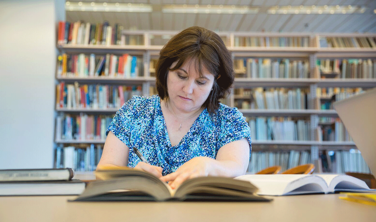 A woman in a blue patterned blouse sits at a library table, focused on taking notes from an open book. Shelves filled with books are visible in the background, creating a scholarly atmosphere.