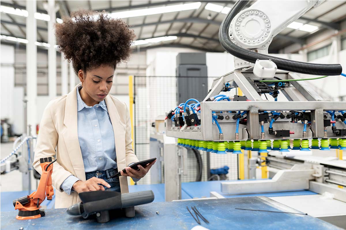 A woman with curly hair stands in a manufacturing facility, using a tablet. She is beside a large industrial machine with numerous wires and suction cups. She appears focused on her work in this modern, high-tech environment.