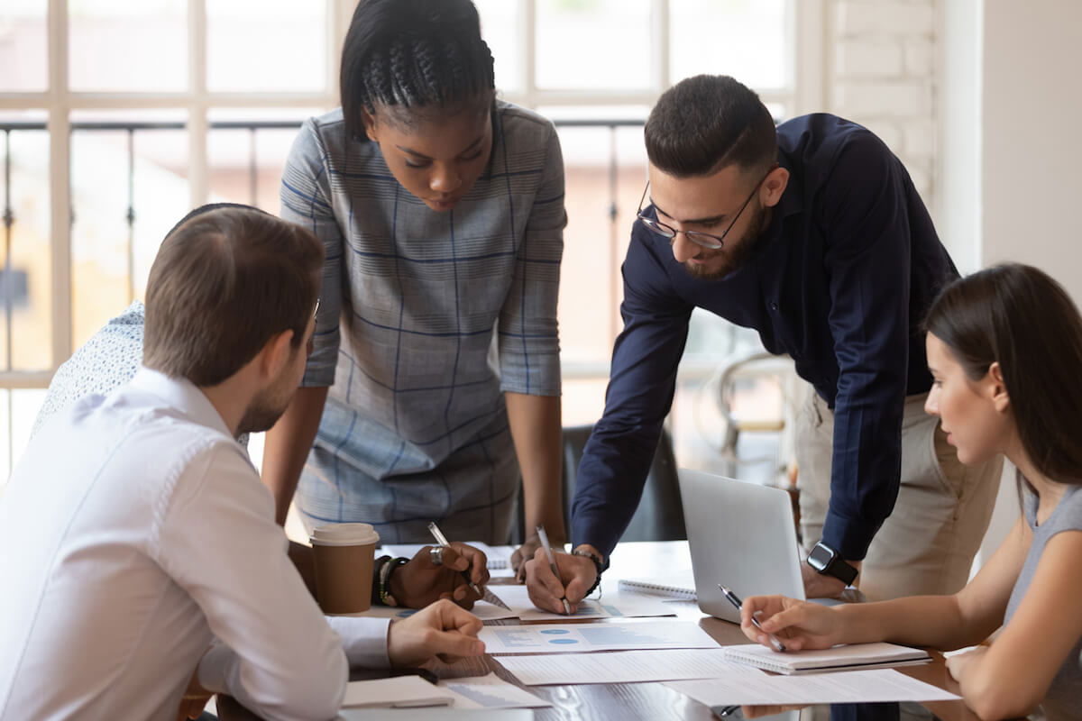 A group of people in an office setting are gathered around a table, intently discussing documents and a laptop. They appear focused and engaged in collaboration.