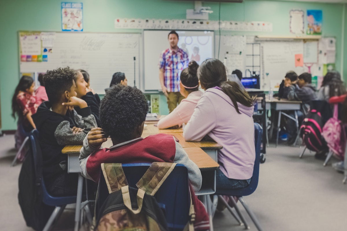 A classroom of students attentively listening to a teacher at the front. Desks are arranged in clusters, and students are taking notes. The teacher stands by a whiteboard with a projector displaying an image. Bags and educational materials are visible.
