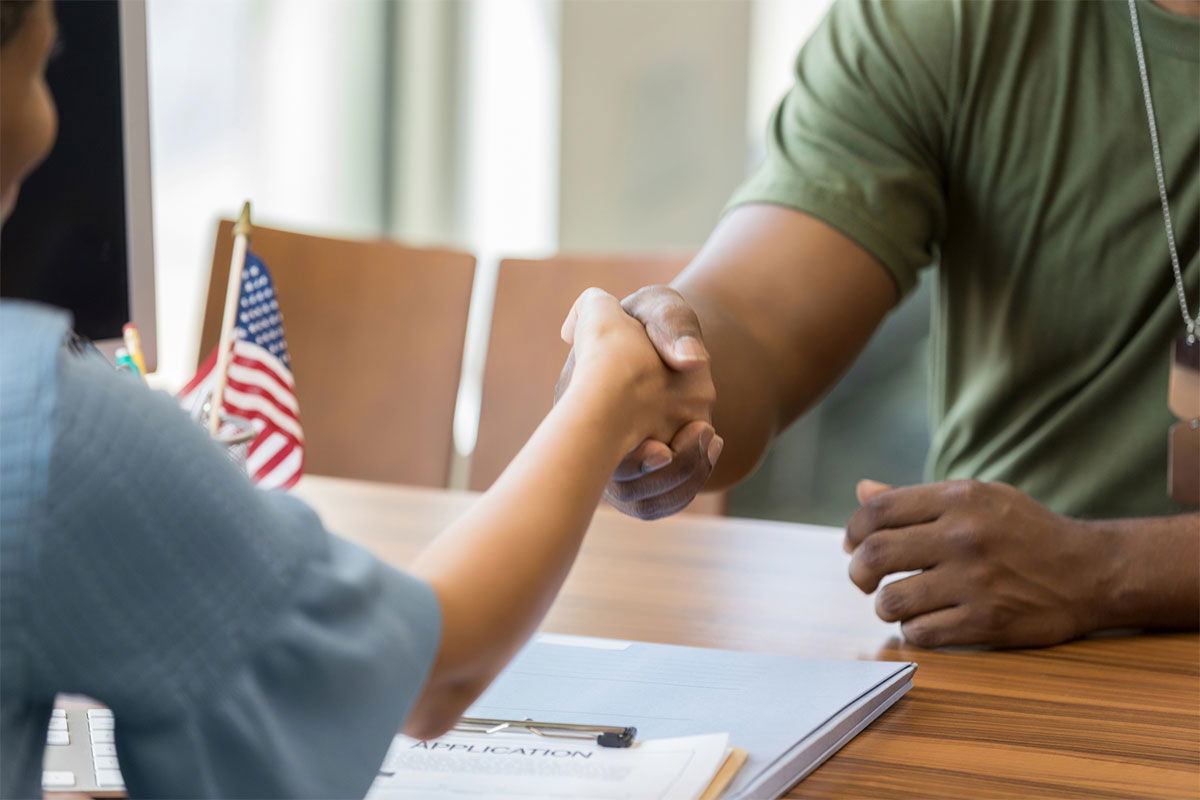 A person in a green shirt and dog tags shakes hands with someone across a table. On the table, there's a small American flag and papers labeled "Application.