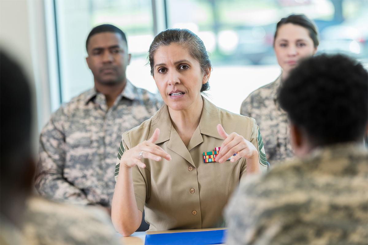 A woman in military uniform leads a discussion with a group of service members in camouflage uniforms. She gestures with her hands, speaking intently. The background is softly blurred, showing windows and natural light.