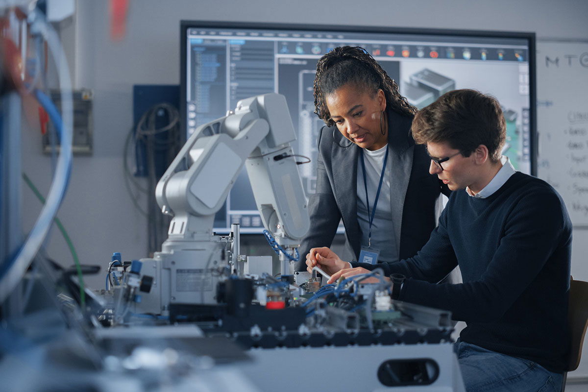 Two people work together in a modern lab, focusing on a robotic arm and electronic components. The background shows computer screens with technical diagrams, indicating an engineering or technology setting.