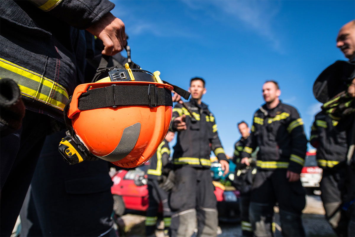 A group of firefighters in uniform stand together outdoors under a clear blue sky. One person is holding an orange helmet. A red fire truck is partially visible in the background.