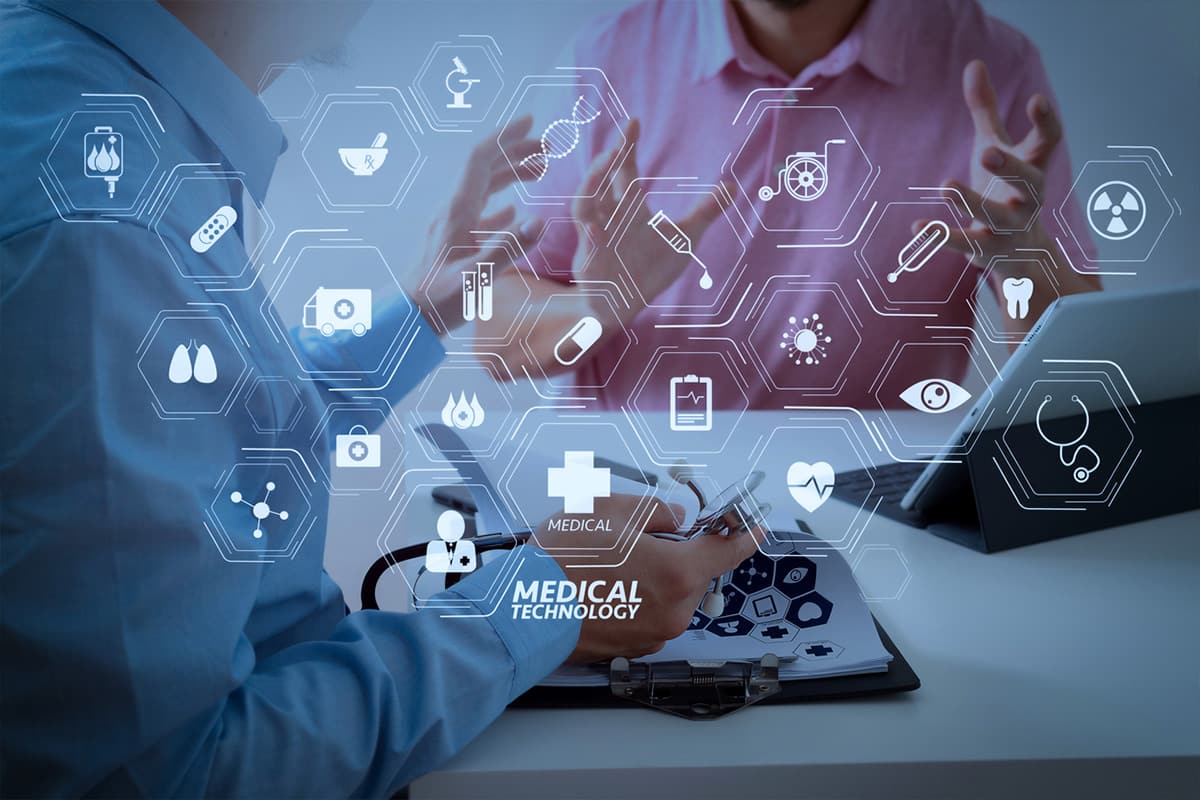 Two people are discussing medical technology at a desk. Various medical icons like a DNA strand, heart, and stethoscope are digitally superimposed over the image, representing healthcare innovation and technological advancement.