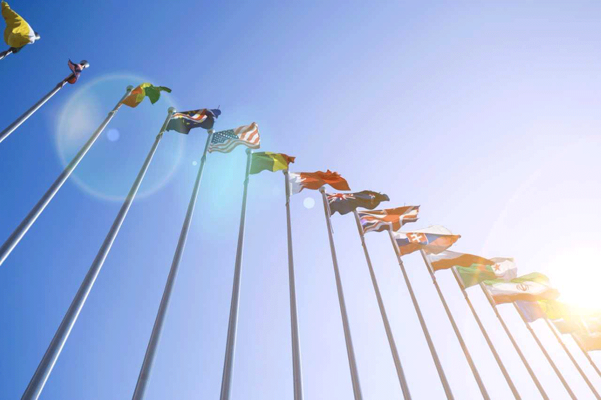 A row of various international flags waving on tall poles against a clear blue sky. The sun shines brightly in the corner, creating lens flare effects.