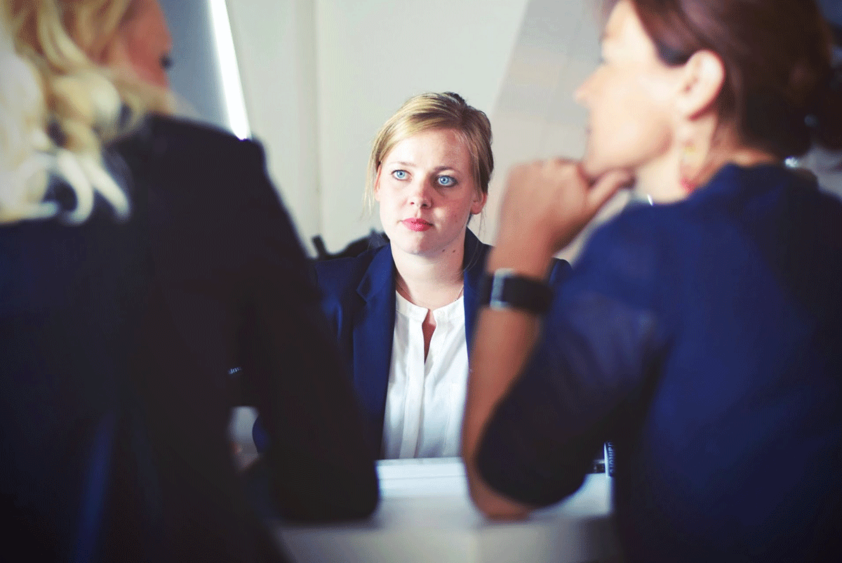 Three people in a professional setting are having a discussion. The person in the center, wearing a dark suit, listens attentively, while the others are seen from behind, focused on the conversation. The atmosphere appears formal and engaged.