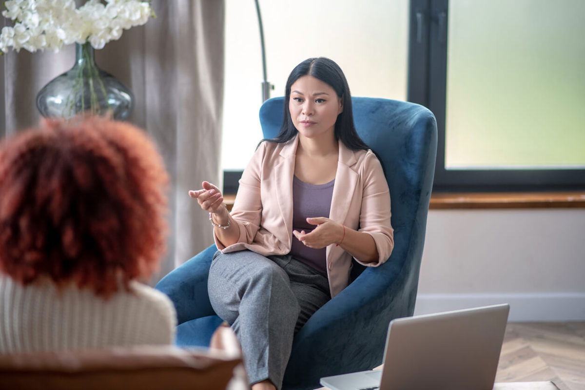 A woman with long dark hair is sitting in a blue armchair, gesturing while speaking. She is wearing a pink blazer and gray pants. Another person with curly red hair is sitting across from her. A laptop is open nearby.
