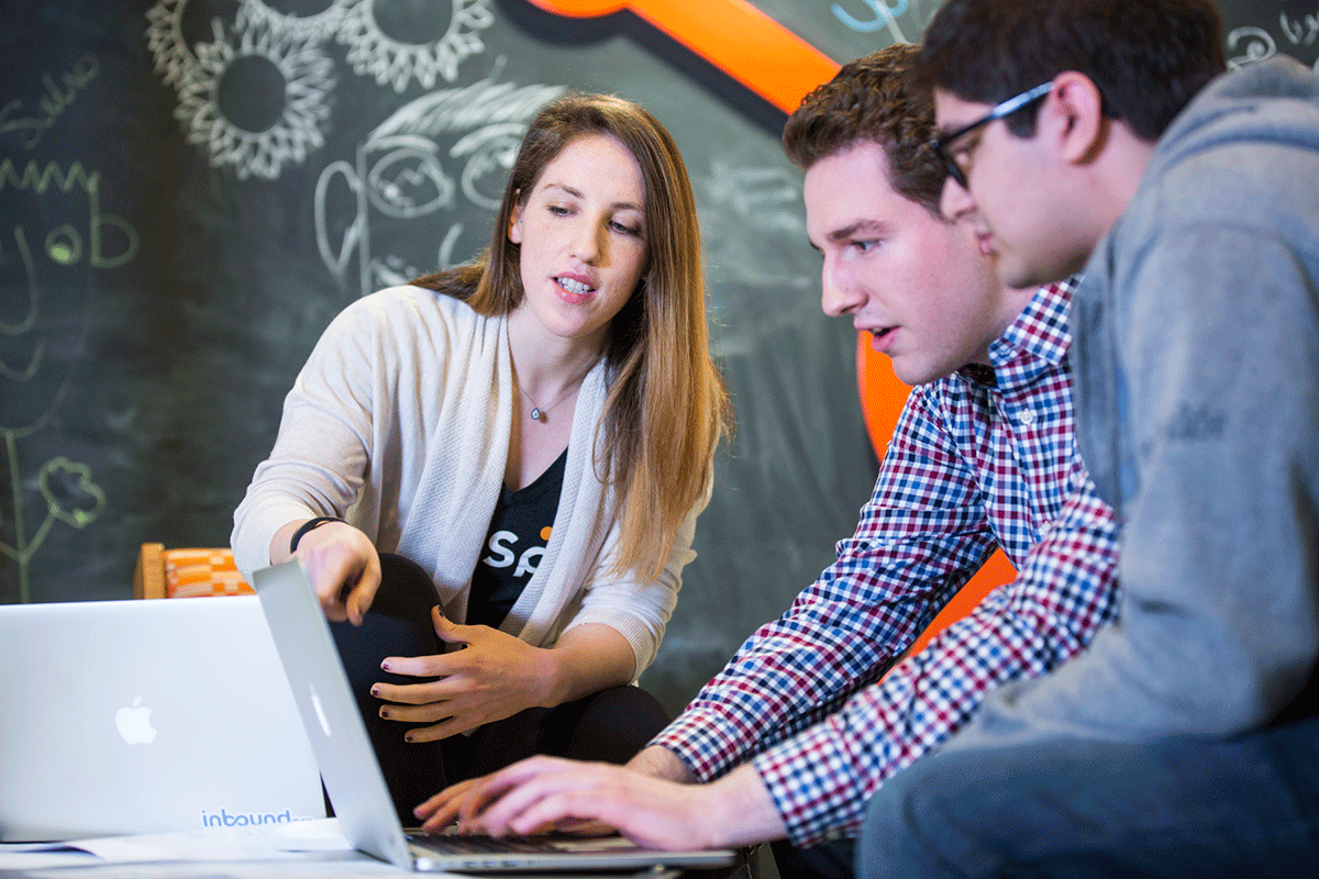 Three people are seated around a laptop, engaged in discussion. One is gesturing towards the screen. They are in a room with a chalkboard wall covered in doodles and writing. The setting appears collaborative and informal.