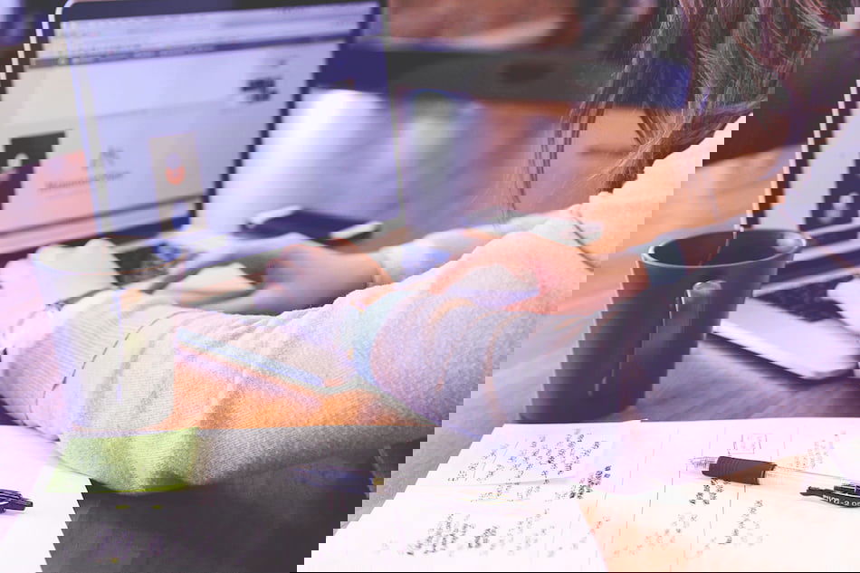 A person typing on a laptop with a webpage open. A notebook, pen, and sticky notes with handwritten text are on the wooden table. A black coffee mug is nearby. The background is softly blurred.