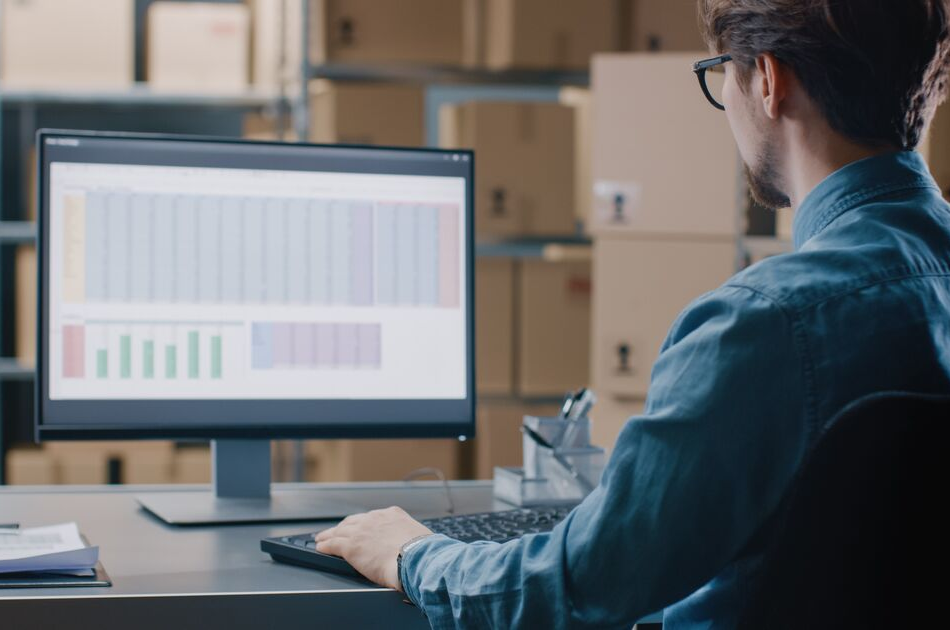 A person in a denim shirt sits at a desk working on a computer. The screen displays a spreadsheet with charts and data. Shelves with boxes are visible in the background.