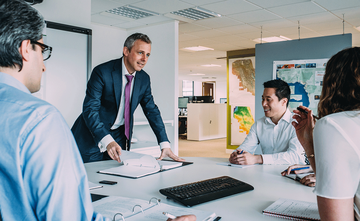 A group of professionals in an office setting engaged in a meeting. A man in a suit stands at the table with documents, while others sit around. A computer keyboard is visible on the table. Wall maps are displayed in the background.